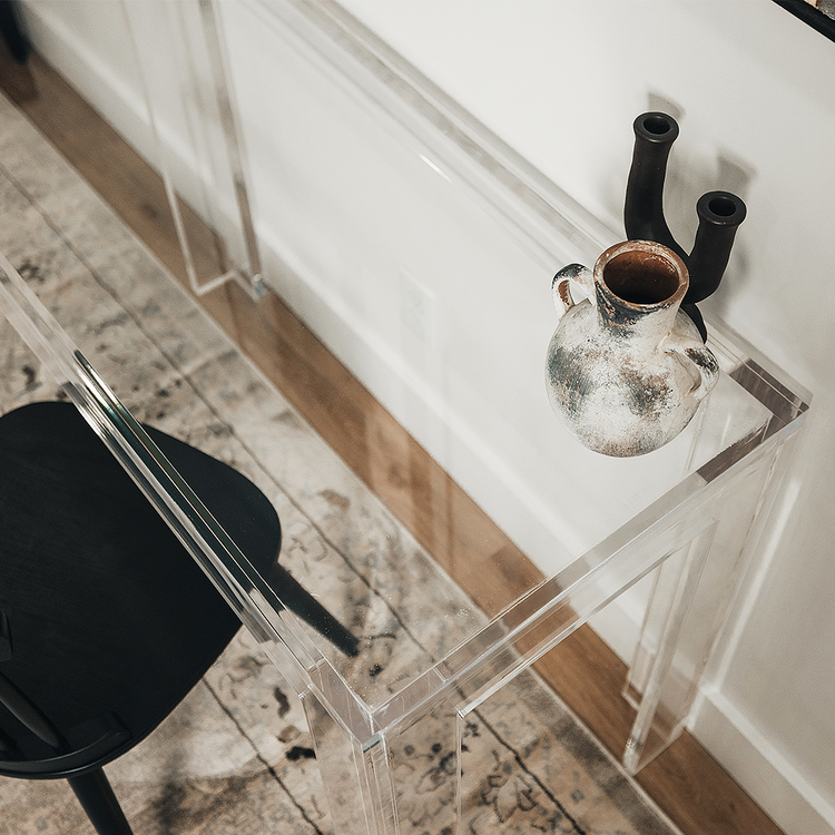 A closeup of the edges of a clear acrylic lucite square leg desk placed against a wall decorated with a vase and candle holder next to a chair. 