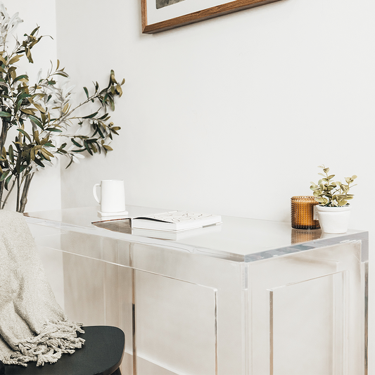 Clear acrylic lucite square leg desk placed against a wall decorated with a candle, plant, coffee cup and a book next to a chair layered with a blanket on the back.  