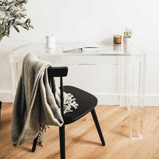 Clear acrylic lucite square leg desk placed against a wall decorated with a candle, plant, coffee cup and a book next to a chair layered with a blanket on the back.  
