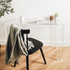 Clear acrylic lucite square leg desk placed against a wall decorated with a candle, plant, coffee cup and a book next to a chair layered with a blanket on the back.  