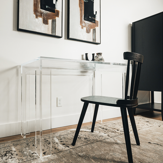 A clear acrylic lucite square leg desk placed against a wall decorated with a vase and candle holder next to a chair. 