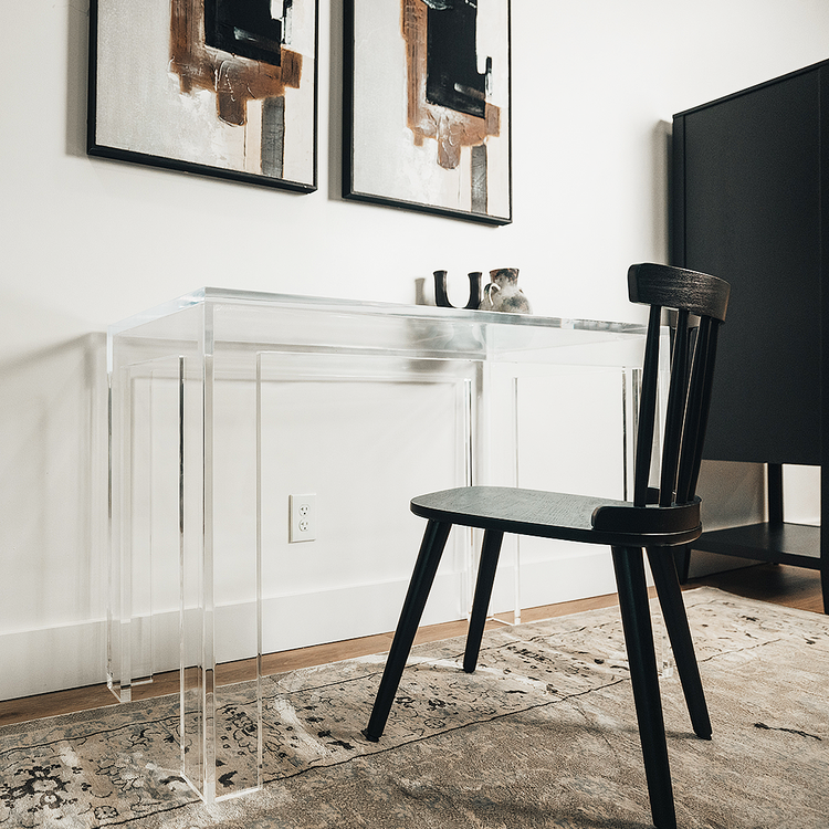 A clear acrylic lucite square leg desk placed against a wall decorated with a vase and candle holder next to a chair. 