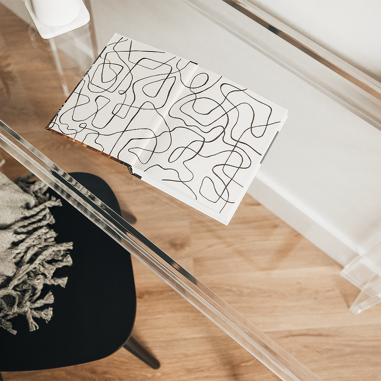 Clear acrylic lucite square leg desk placed against a wall decorated with a coffee cup and a book next to a chair layered with a blanket on the back.  