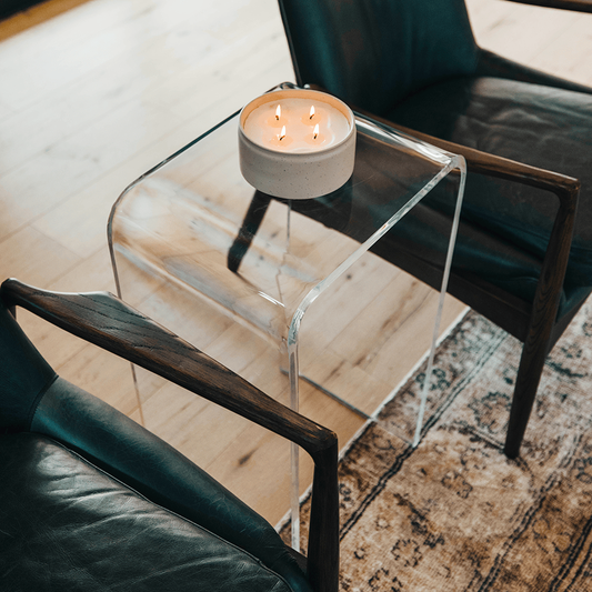 A closeup of a clear acrylic lucite waterfall edge side table with a candle on top placed between 2 leather chairs in a living room setting. 