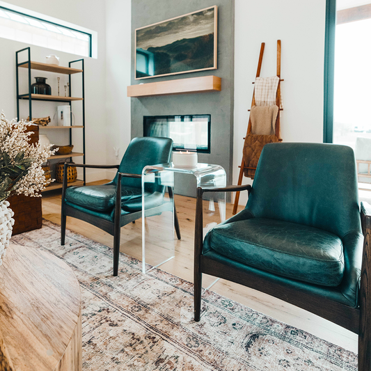 A clear acrylic lucite waterfall edge side table with a candle on top placed between 2 leather chairs in a living room setting. 
