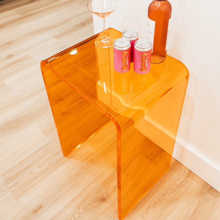 A orange tinted acrylic lucite waterfall edge side table with a wine bottle, soda, and a wine glass against a wall in a kitchen. 