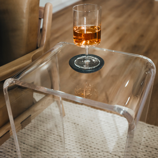 A closeup of the edges of a clear acrylic lucite waterfall end table holding a wine glass next to an easy chair. 