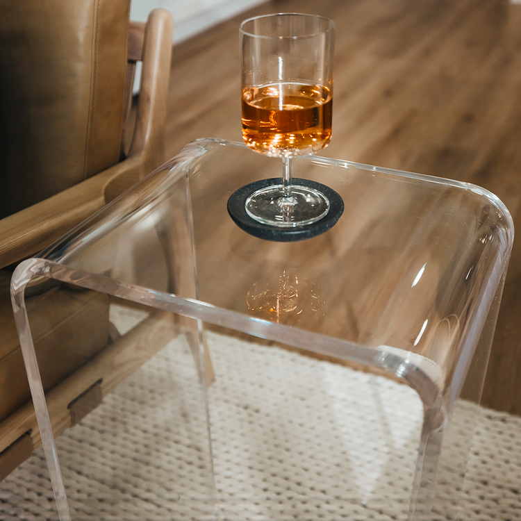 A closeup of the edges of a clear acrylic lucite waterfall end table holding a wine glass next to an easy chair. 