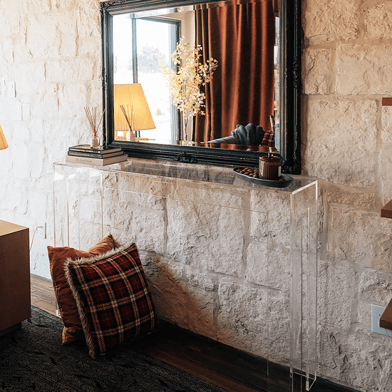 Modern clear acrylic console table against a white stone wall with a mirror above and decorative items placed in a living room setting.