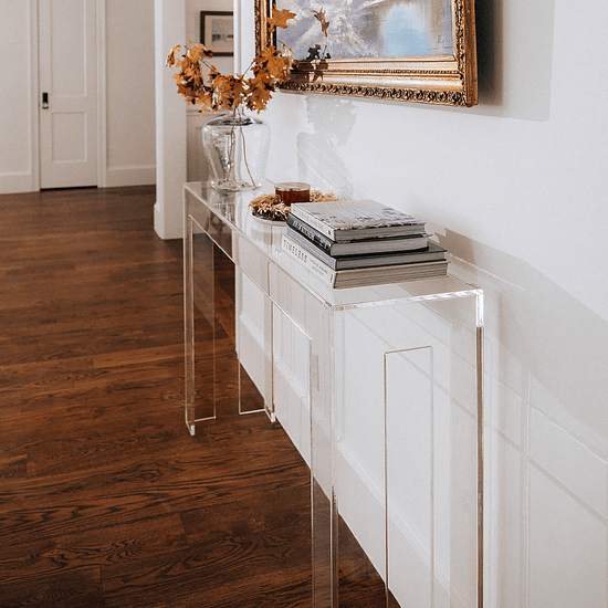 A side view of a modern clear acrylic console table against a white wall with decorative items placed on a wooden floor of a hallway.