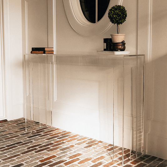 Modern clear acrylic console table against a white wall with decorative items placed in a brick floor foyer.