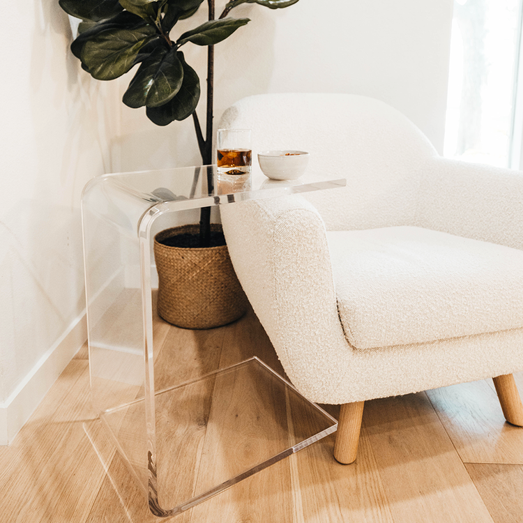 A C-Shaped acrylic lucite side table with a beverage glass and snacks on top partially slid underneath an easy chair in a corner of a living room next to a window and a plant.  