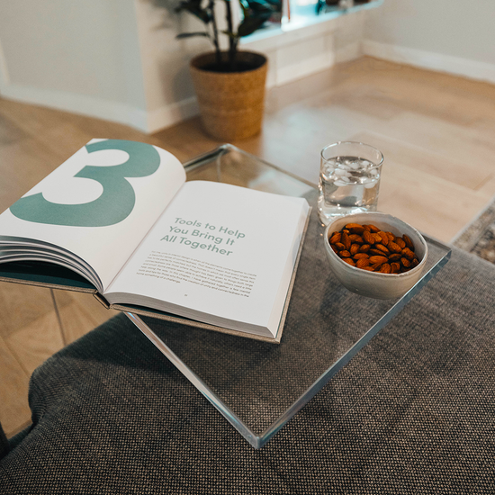 A C-Shaped acrylic lucite side table with a beverage glass, snacks, and a book on top partially slid underneath a sofa in a living room. 