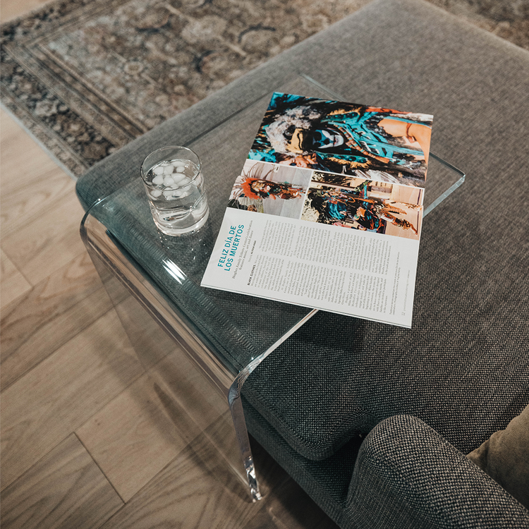 A C-Shaped acrylic lucite side table with a beverage glass and a book on top partially slid underneath a sofa in a living room. 