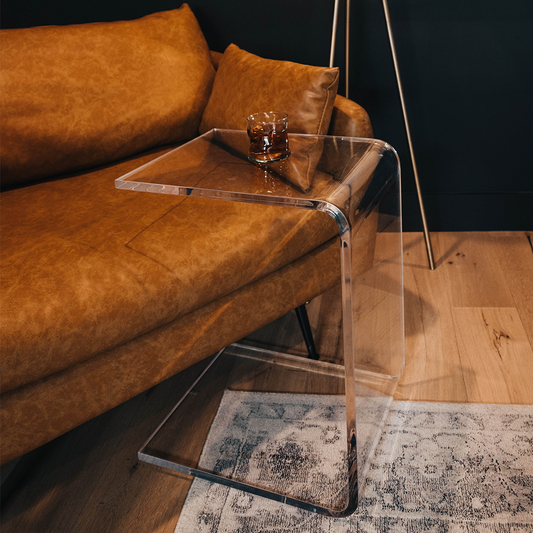 A C-Shaped acrylic lucite side table with a beverage glass on top partially slid underneath a brown leather sofa. 