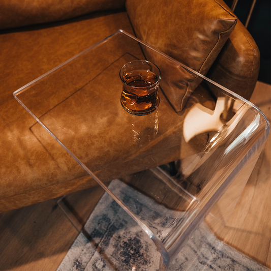 A C-Shaped acrylic lucite side table with a beverage glass on top partially slid underneath a brown leather sofa. 
