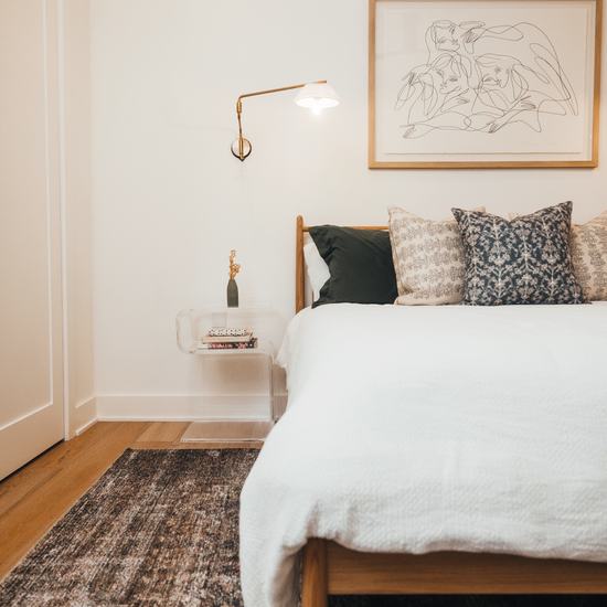 A clear acrylic lucite side table in a modern s-shape with a small vase of flowers on top and books on the interior shelf placed beside a bed.