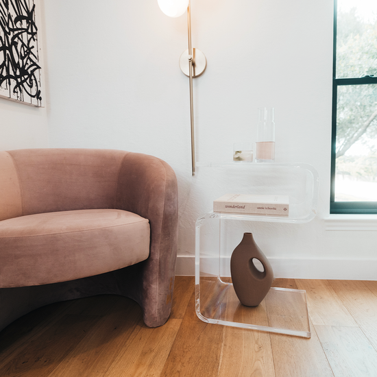 A clear acrylic lucite side table in a modern s-shape with drinks on top and a book on the interior shelf placed beside an easy chair in a living room next to a window and a lamp. 