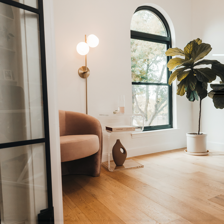 A clear acrylic lucite side table in a modern s-shape with drinks on top and a book on the interior shelf placed beside an easy chair in a living room next to a window and a lamp. 