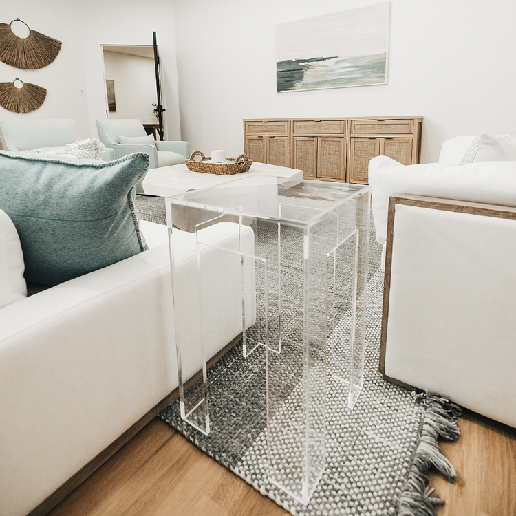 A clear acrylic side table with a square leg style base placed between a white couch and a white chair in a modern living room.