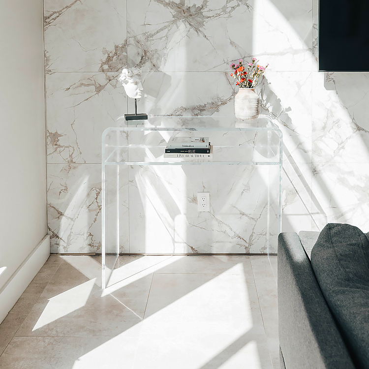 A clear acrylic waterfall console table with 1 interior shelf decorated with a vase of flowers, books, and a bust statue placed in a brightly lit modern living room next to a marble tiled wall. 
