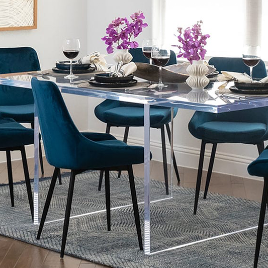 Clear acrylic slab style dining room table displaying centerpieces of flower vases, wine glasses, and place settings surrounded by chairs on an area rug in a dining room.