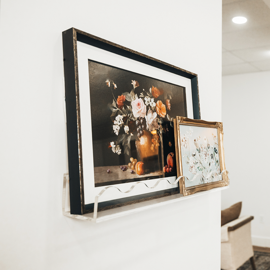 Framed floral artwork on a white wall being held by a clear acrylic floating bookshelf with a wavy edge. 