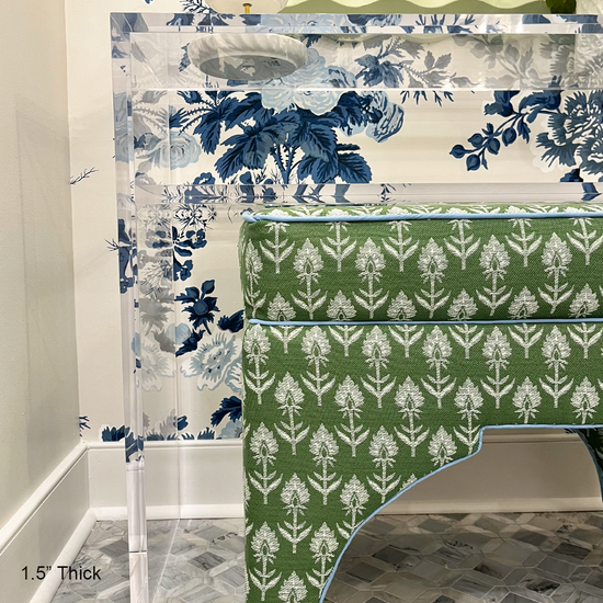 A closeup of a clear acrylic console table with 1 interior shelf decorated with a plant and a jewelry dish placed against a wall with floral wallpaper and a mirror. An upholstered vanity bench is placed underneath. 