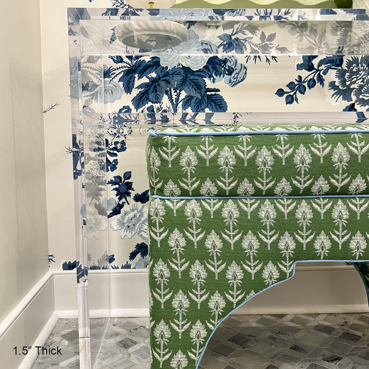 A closeup of a clear acrylic console table with 1 interior shelf decorated with a plant and a jewelry dish placed against a wall with floral wallpaper and a mirror. An upholstered vanity bench is placed underneath. 