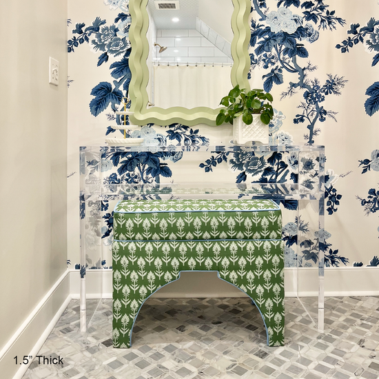 A clear acrylic console table with 1 interior shelf decorated with a plant and a jewelry dish placed against a wall with floral wallpaper and a mirror. An upholstered vanity bench is placed underneath. 
