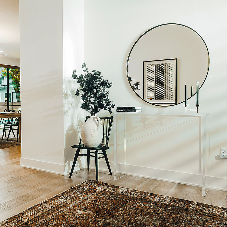 A clear acrylic console table decorated with books and candles placed against a wall with a mirror in the entryway of a home. 
