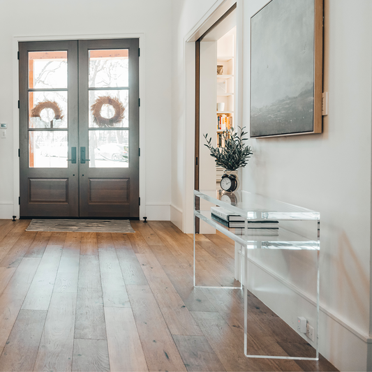 A clear acrylic console table with 1 interior shelf decorated with a plant and a clock placed against a wall with framed artwork in the entryway of a home. 