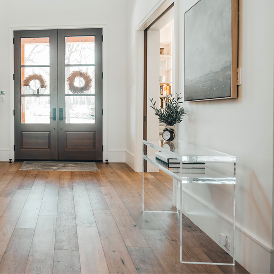A clear acrylic console table with 1 interior shelf decorated with a plant and a clock placed against a wall with framed artwork in the entryway of a home. 