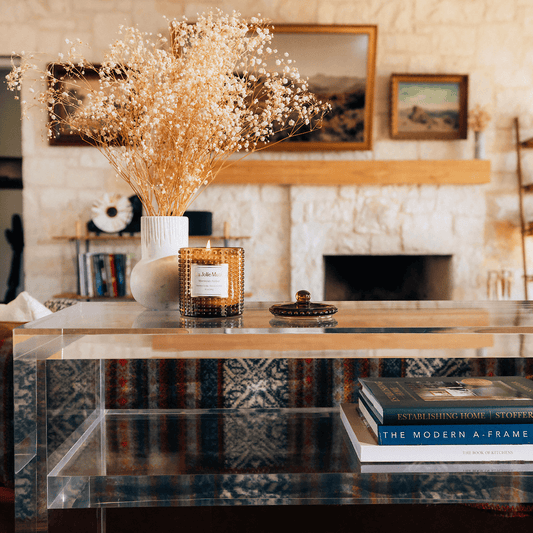 A closeup of the thick edges on a clear acrylic console table with books on the interior shelf and decor on top placed behind a couch in a living room setting.