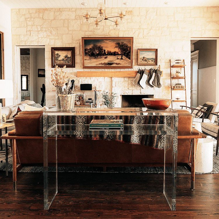 A closeup of the thick edges on a clear acrylic console table with books on the interior shelf and decor on top placed behind a couch in a living room setting.