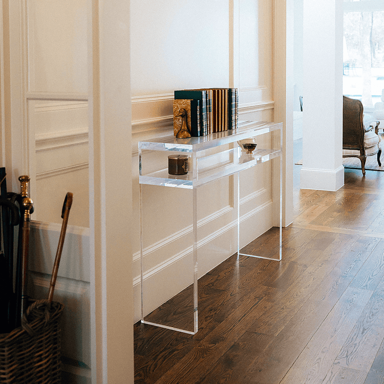A clear acrylic console table with decor on the interior shelf and books on top against a wall in an interior foyer area.