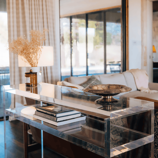 A closeup of the thick edges on a clear acrylic console table with books on the interior shelf and decor on top placed behind a couch in a living room setting.
