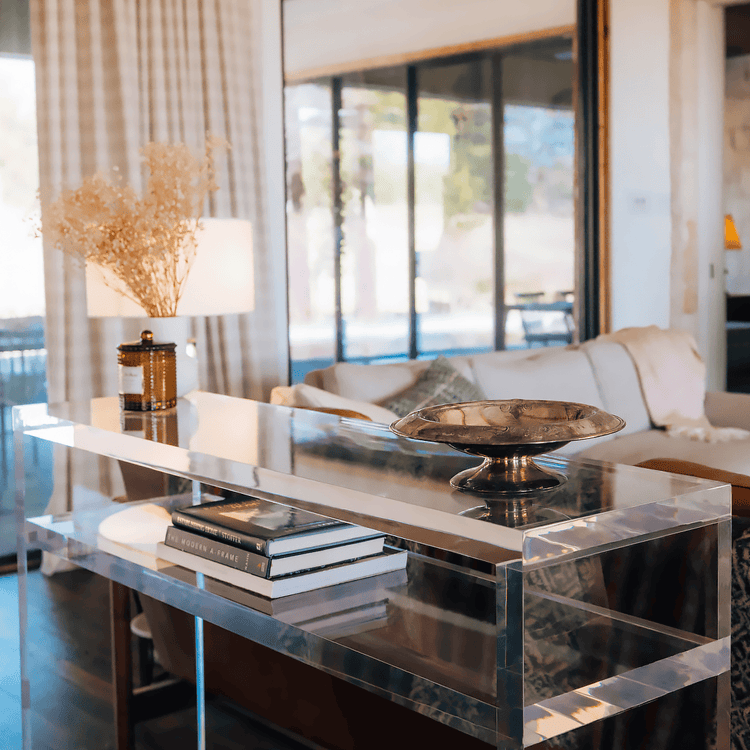 A closeup of the thick edges on a clear acrylic console table with books on the interior shelf and decor on top placed behind a couch in a living room setting.