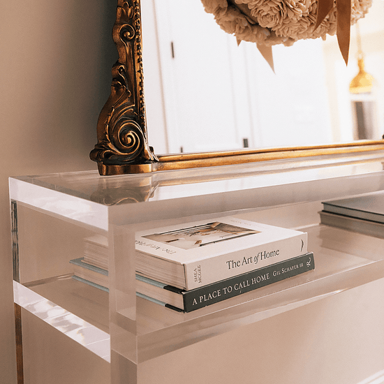 A closeup of the thick edges on a clear acrylic console table with books on the interior shelf and a mirror on top against a wall in an interior foyer area.
