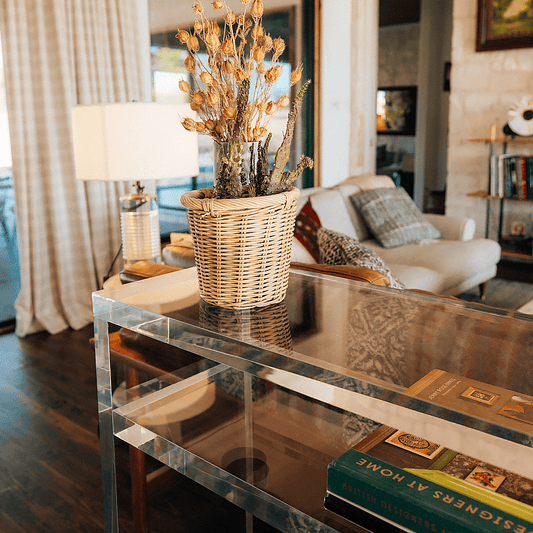 A closeup of the thick edges on a clear acrylic console table with books on the interior shelf and decor on top placed behind a couch in a living room setting.