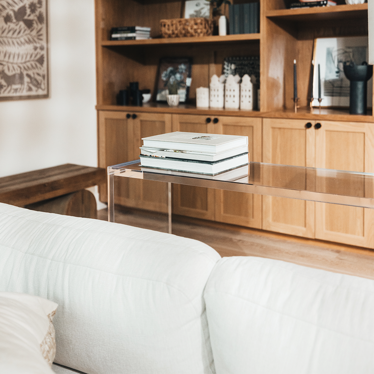 A modern clear acrylic console table with a slab style reflective surface, placed in a room with a beige sofa and books on top.