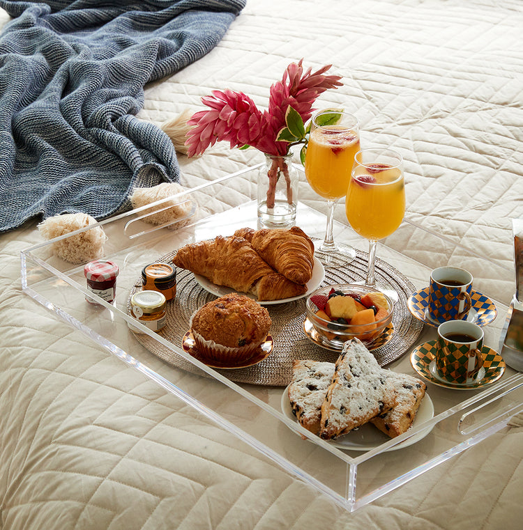 Clear acrylic serving tray with sides on a bed displaying a small vase of fresh cut flowers, 2 stemmed orange juice glasses, plates of muffins, scones, fruit and 2 cups of coffee.