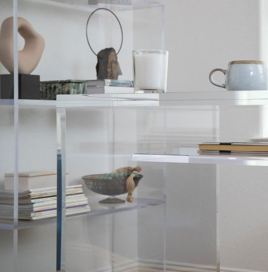 Closeup of a clear acrylic slab style desk with 2 interior shelves on the left and right sides of the desk displaying books, candle, and coffee cup. Also featured is a clear acrylic tall bookcase.