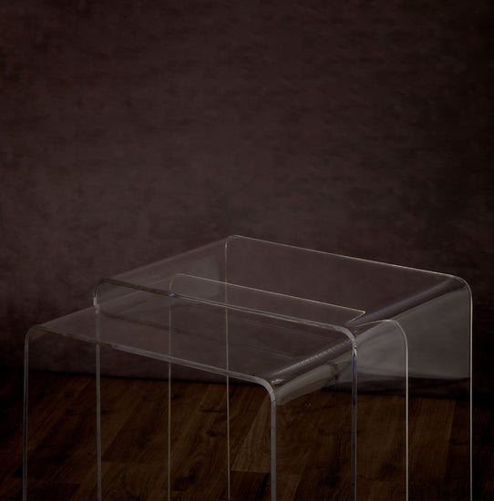 Catalog closeup of 2 clear acrylic waterfall edge nesting end tables on a hardwood floor with lower table pulled out.
