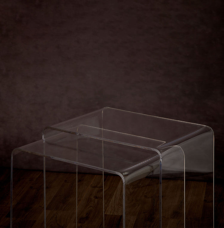 Catalog closeup of 2 clear acrylic waterfall edge nesting end tables on a hardwood floor with lower table pulled out.