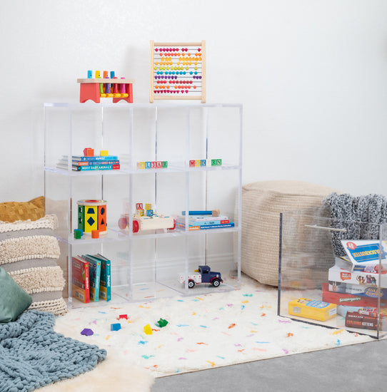 Clear acrylic 9 cube storage unit displaying children's toys, games, and books in a playroom furnished with pillows and an area rug. The unit features 3 rows of 3 cubes.