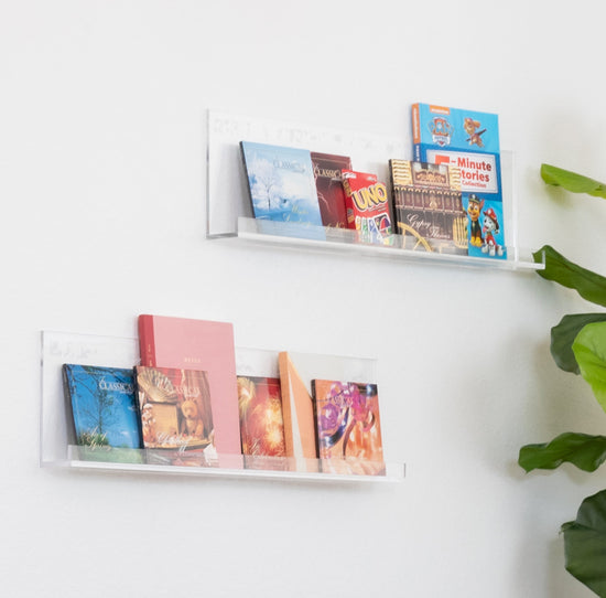 A set of 2 clear acrylic floating bookshelves displaying books mounted on a wall.