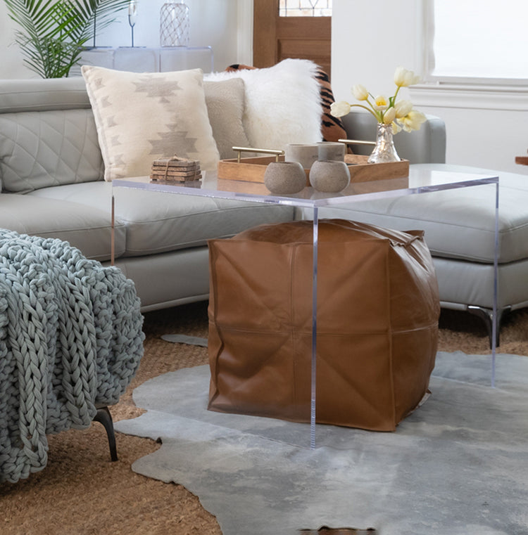 Clear acrylic 23" slab coffee table, with a pouf ottoman slid underneath, displaying vases, coasters, and a serving tray placed on a fur rug in front of a couch in a living room.