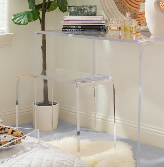 Clear acrylic waterfall edge vanity stool placed under a clear acrylic waterfall edge vanity displaying books, perfumes, and art decor in a bedroom.