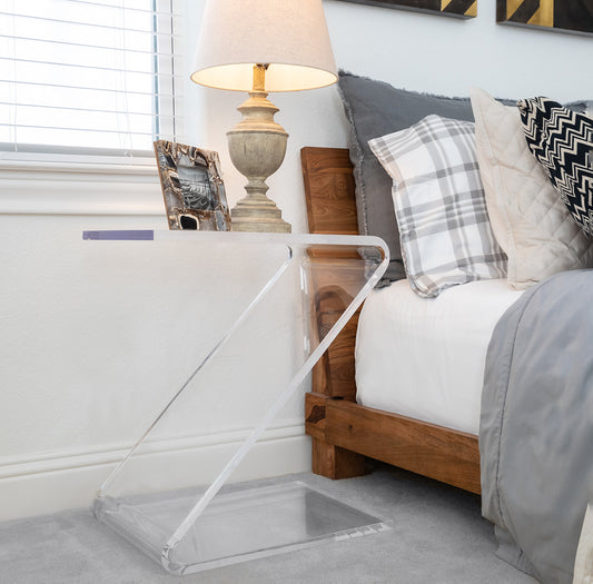 Clear acrylic waterfall edge z shaped end table displaying a lamp and photo frame next to a bed.